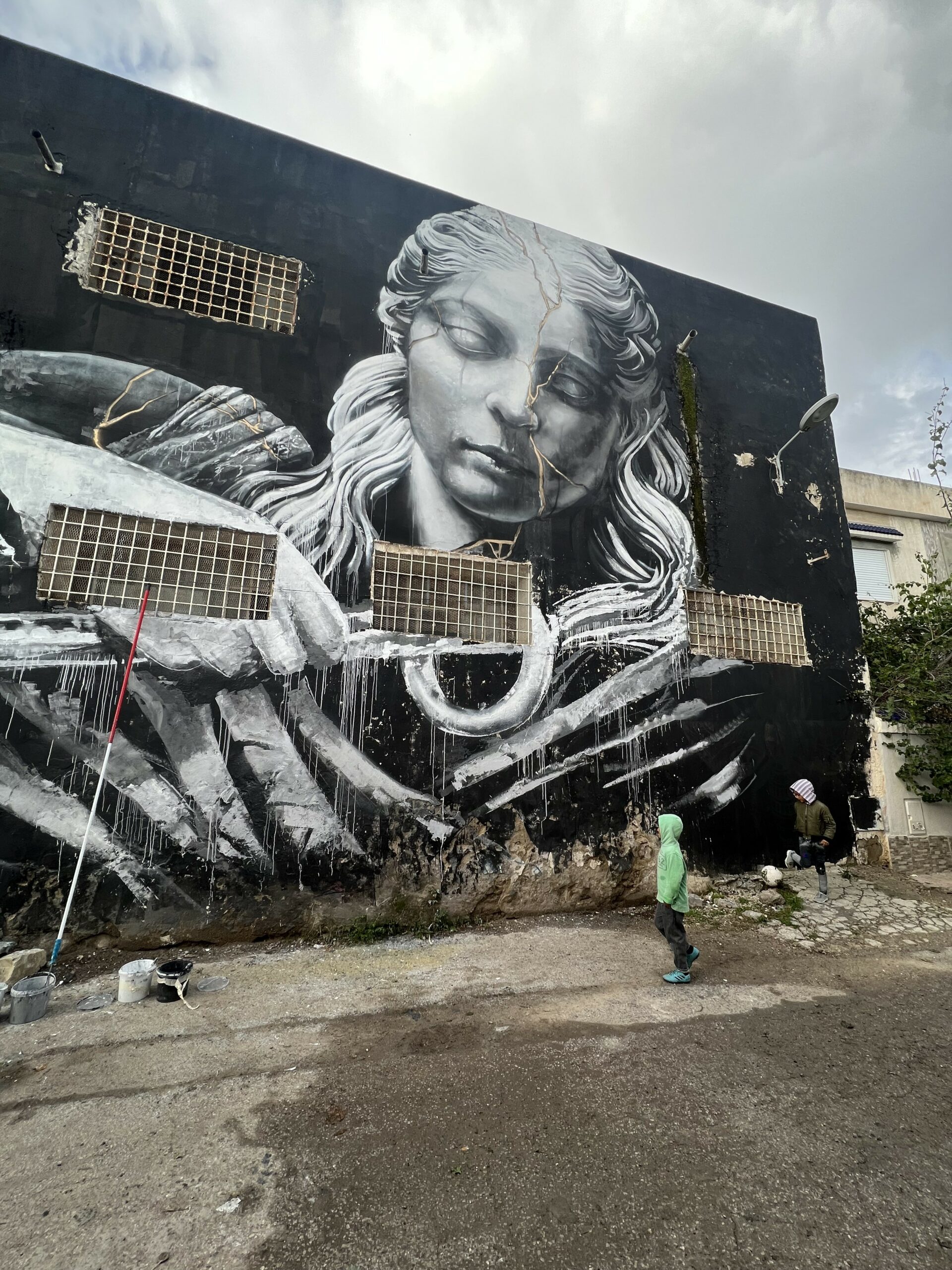 "HOPE" by Neve, Tunis 2023 — in-progress view; mural largely complete; classical figure emerging in grey-white from black wall; industrial building with metal grates visible; figure in background; Tunisia February 2023